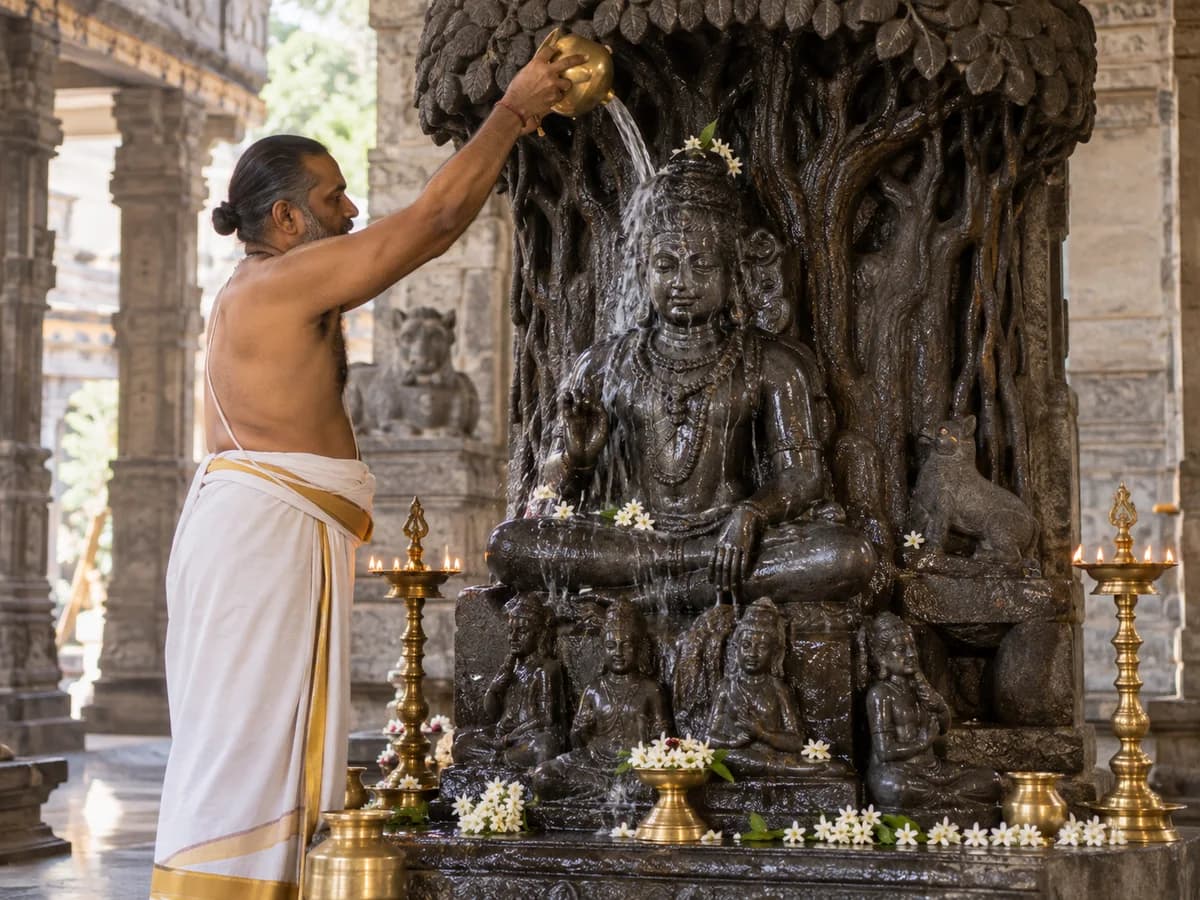 A priest pours water over a traditional Dakshinamurthy idol adorned with flowers beside brass lamps.