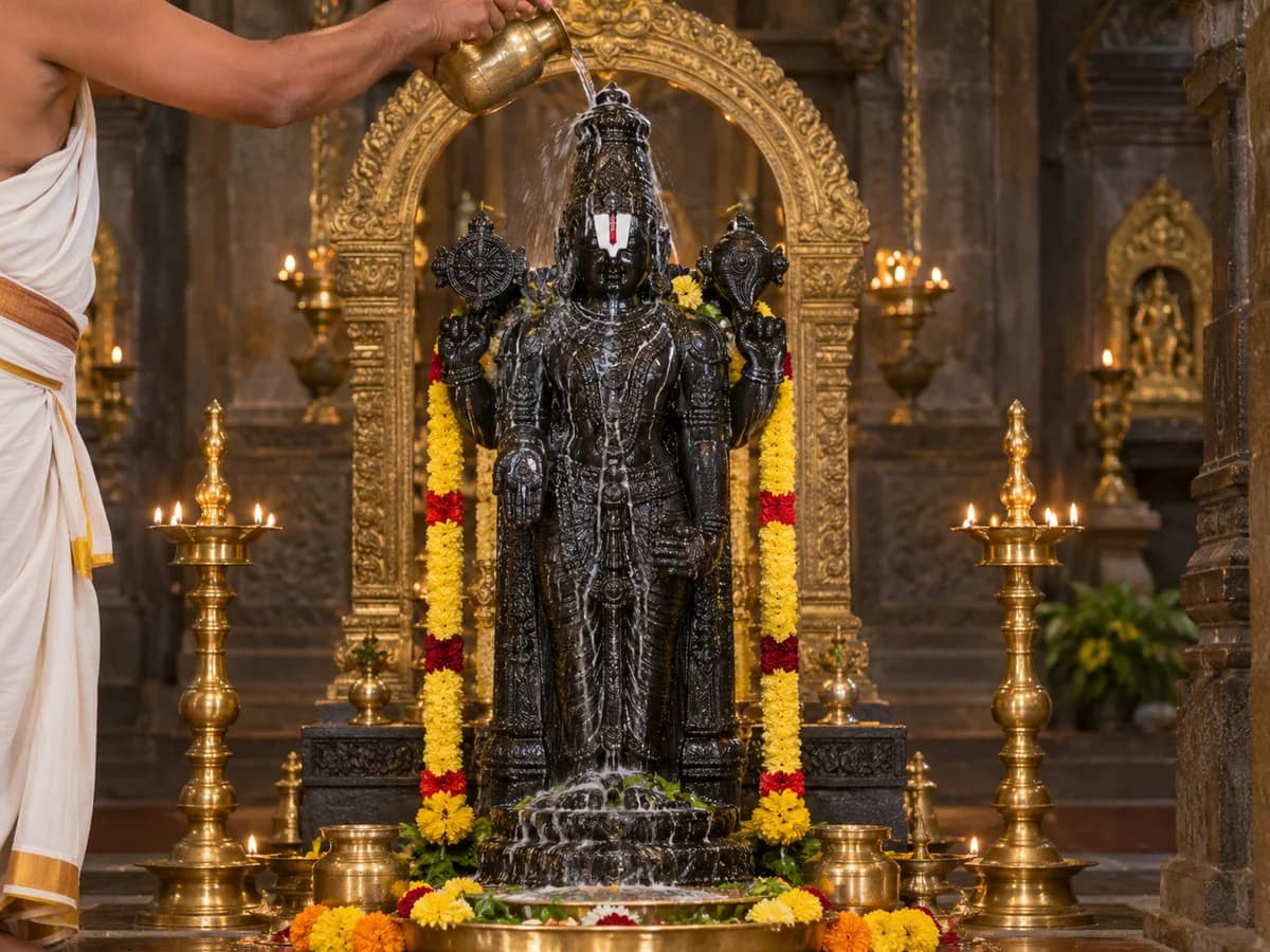 A priest pours water over a Venkateswara idol in a temple shrine with brass lamps and flowers.